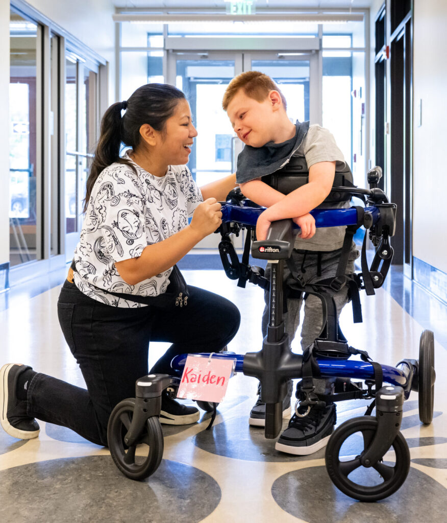 Physical Therapist kneels next to student who is using a walking device. They look at each other smiling in a hallway.