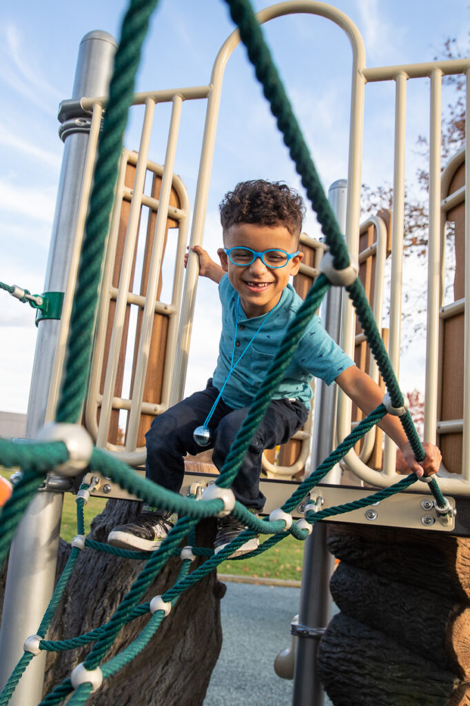 MSB student, Ein, wearing blue glasses and a blue shirt climbs on a playground rope bridge, smiling.