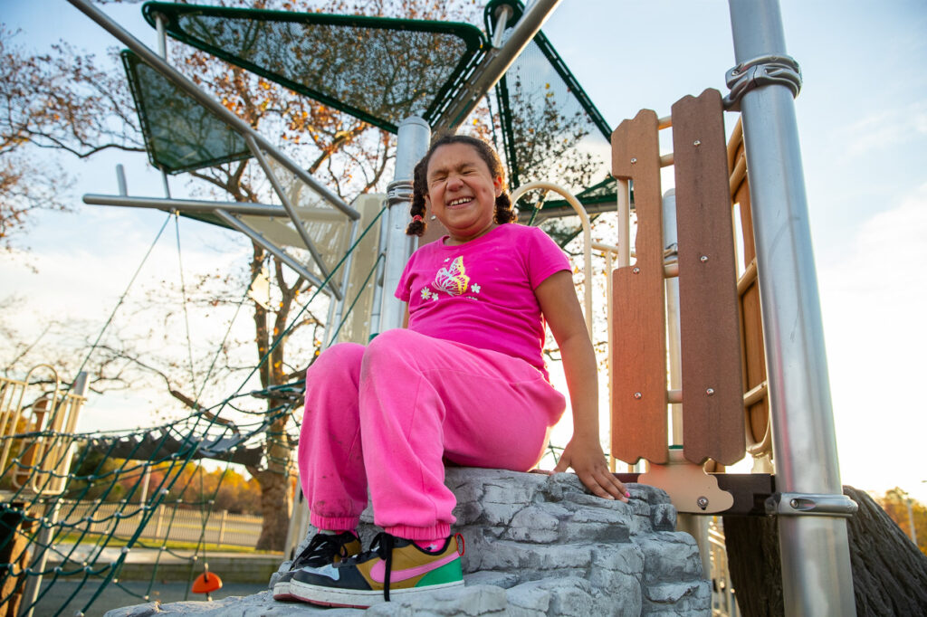 MSB Student, Ren, in a pink outfit sits smiling on playground equipment. Trees and a clear sky are in the background.