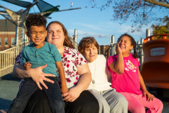 A family of four sitting together on a sunny day, with a playground structure in the background. All are smiling.