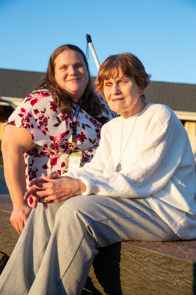 A mother and daughter (Melissa & Dorothy) sitting outdoors on a sunny day, one wearing a floral blouse and smiling, and the other in a white sweater, looking ahead.