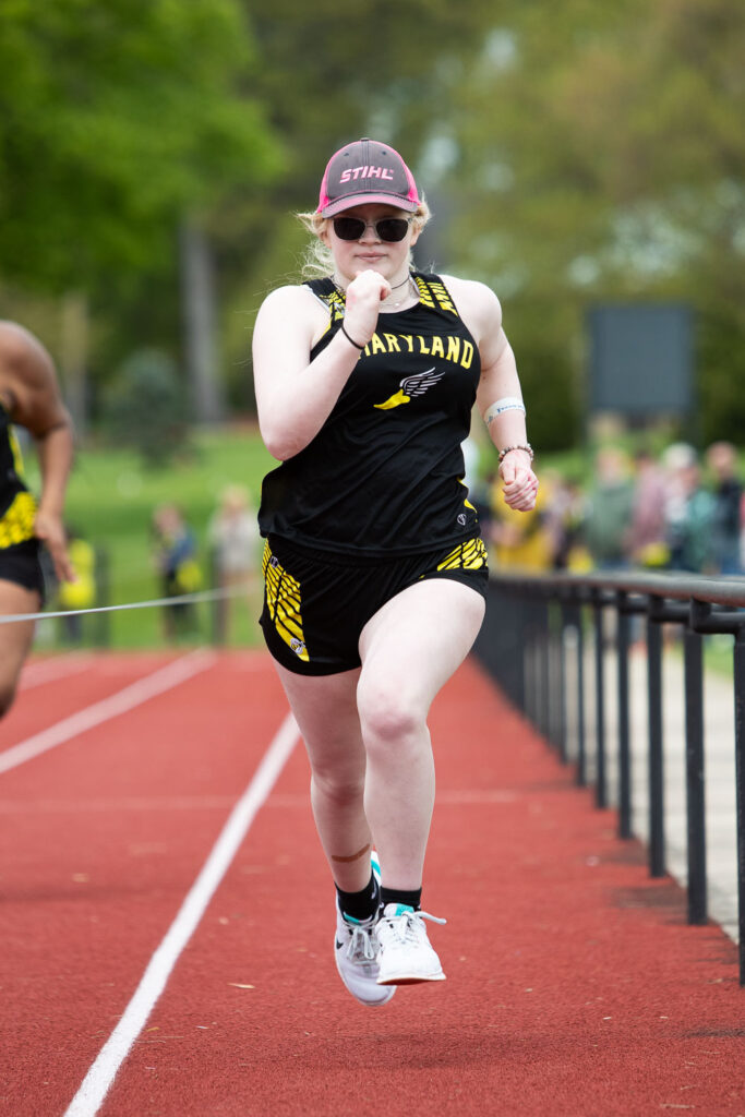 A female athlete in a black and yellow Heartland uniform sprints down a red track, focused ahead. She wears sunglasses, a red cap, and white running shoes. Spectators and greenery are visible in the background.