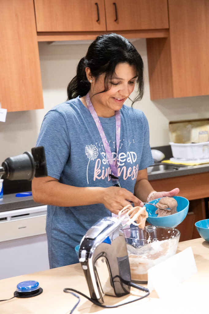 A woman in a blue "Spread Kindness" shirt smiles while scooping cookie dough from a bowl in a kitchen, with a mixer and ingredients on the counter.