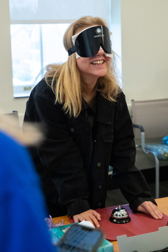 A person with long blonde hair smiles while wearing a black blindfold and standing at a table with their hands on it. A small silver bell and various items are on the table. The background shows chairs and a window.
