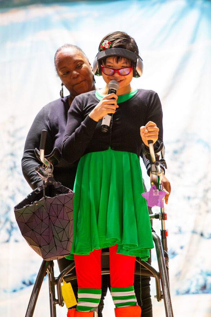 A young person in a green dress, red pants, and headphones stands with a walker, holding a microphone. An adult stands supportively behind them. They are on a stage with a blue and white background.