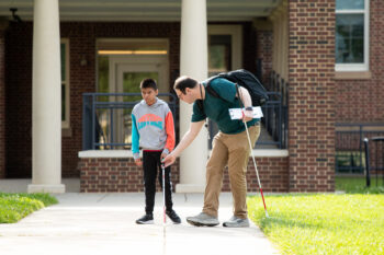 A man is helping a boy with a white cane walk along a sidewalk in front of a brick building. The man points to the ground, carries a backpack, and the boy listens attentively—it's a supportive Back to School moment at MSB.