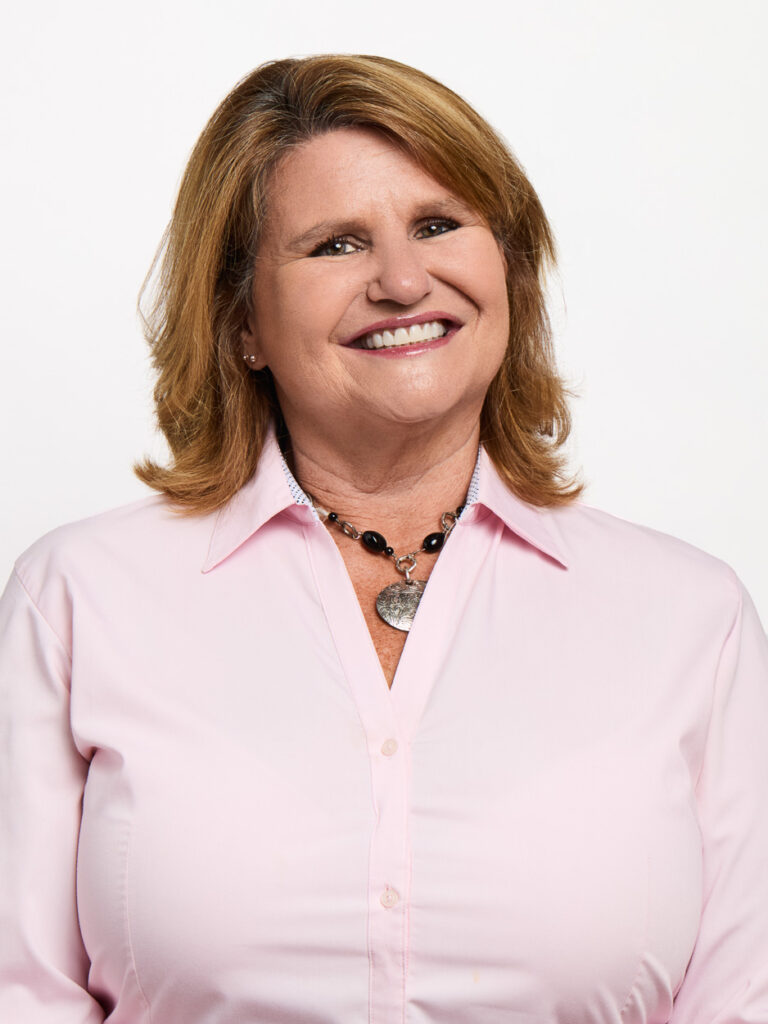 A woman with shoulder-length light brown hair smiles at the camera. She is wearing a light pink button-up shirt and a black beaded necklace with a heart-shaped pendant, representing MSB Leadership against a plain white background.