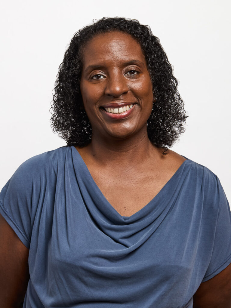 A woman with short curly hair smiles at the camera. She is wearing a blue draped top and exudes confidence, embodying MSB Leadership, as she poses against a plain white background.