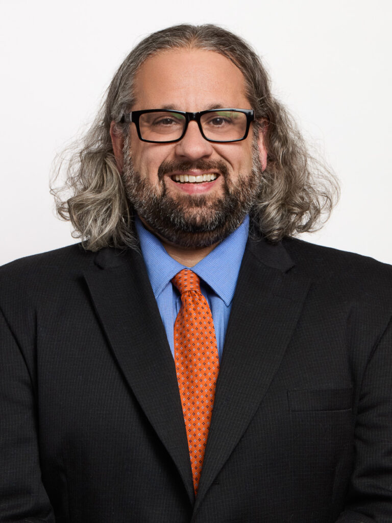 A smiling man with long gray hair, a beard, and glasses wears a black suit, blue shirt, and orange patterned tie, embodying MSB leadership as he stands against a plain white background.