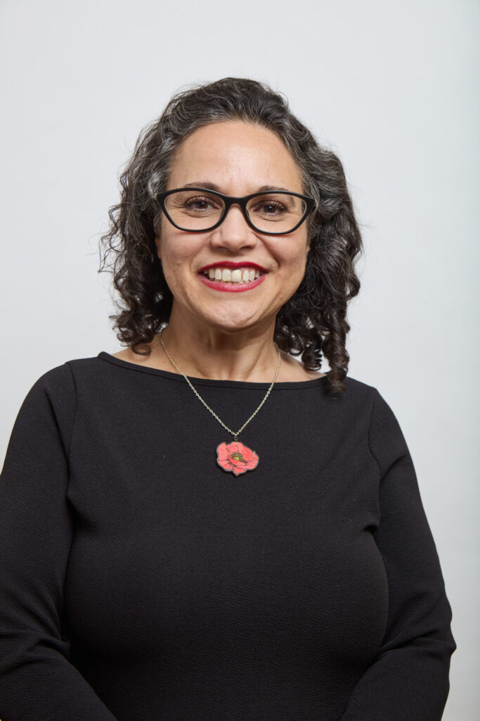 A smiling woman with curly dark hair, wearing glasses, a black top, and a necklace with a red flower pendant, stands in front of a plain white background—radiating confidence and MSB Leadership.