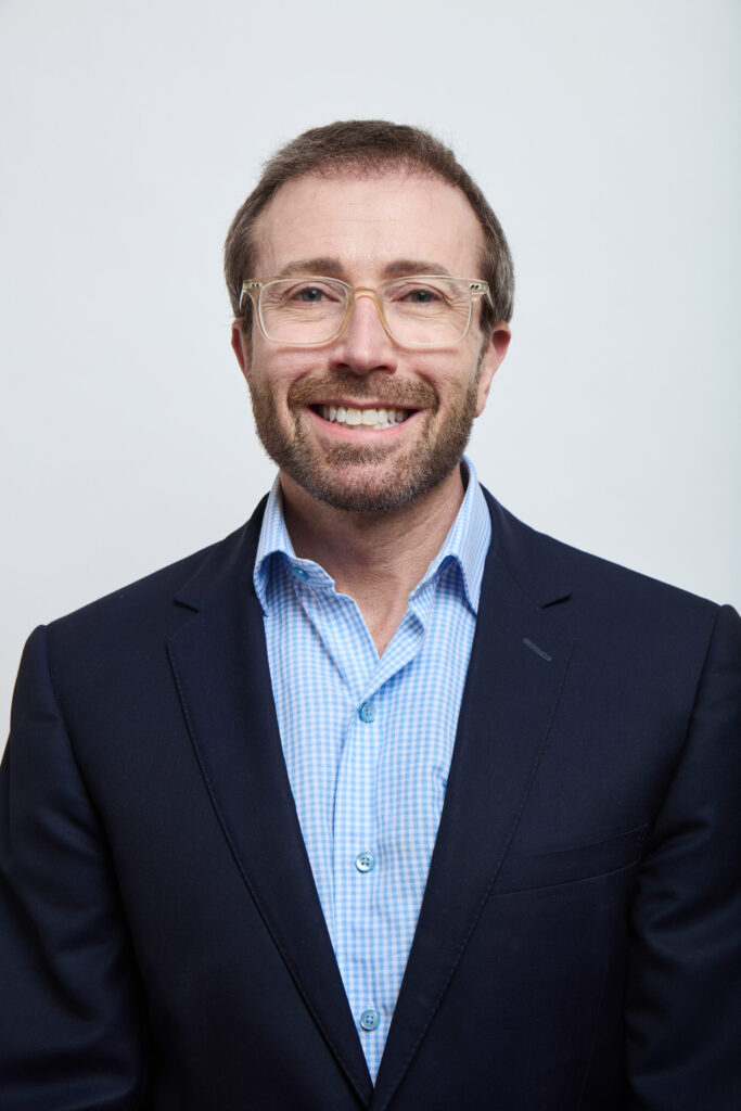 A man with short brown hair, a beard, and glasses is smiling. He is wearing a navy blazer over a light blue checkered dress shirt, standing against a plain white background, reflecting the confidence of MSB Leadership.